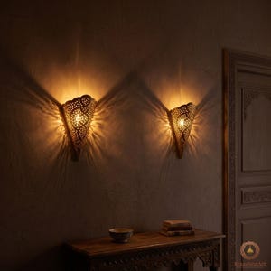 Peut inclure: Deux appliques murales en laiton avec des motifs ajourés complexes, projetant une lumière chaude et structurée sur le mur. Les appliques sont fixées à un mur texturé et sombre, créant une ambiance chaleureuse. Une table en bois avec un bol et des livres est en dessous.