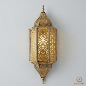 May include: Ornate gold-colored wall sconce with intricate cut-out patterns. The lamp has a domed top and a pointed bottom, with a warm light emanating from within. The design is reminiscent of Middle Eastern or Moroccan style.