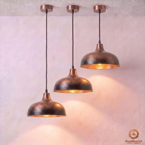 May include: Three pendant lights with hammered copper domes and black cords suspended from the ceiling. Each fixture features a rounded metal shade with a textured exterior and a warm, reflective interior finish, casting a soft downward glow.