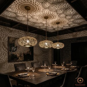 Peut inclure: Trois lampes suspendues en laiton avec des motifs ajourés complexes sont suspendues au-dessus d'une table à manger en bois sombre. Les lampes projettent une lueur à motifs sur le plafond et la table. La pièce a une ambiance chaleureuse et sophistiquée.