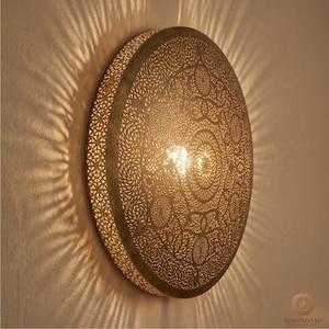 May include: A round, brass-colored wall sconce with intricate, laser-cut patterns. The light shines through the cutouts, casting decorative shadows on the wall. The sconce has a warm, inviting glow, perfect for ambient lighting.