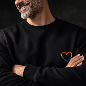May include: Black sweatshirt with a rainbow heart design on the chest. The heart is outlined in a spectrum of colors, including red, orange, yellow, green, blue, and purple. The sweatshirt is made of a soft material.