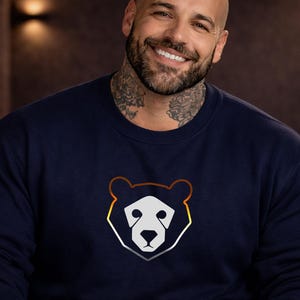 Puede incluir: Sudadera azul marino con un diseño de cara de oso. La cara del oso es blanca, delineada en naranja, amarillo y gris. El gráfico está en la parte delantera de la parte superior de cuello redondo y manga larga.