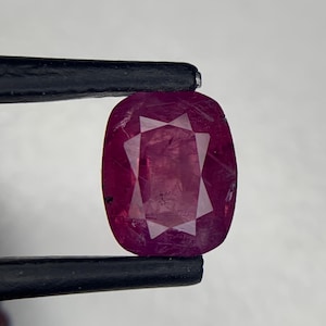 Può includere: Una gemma rosso scuro con taglio a cuscino tenuta da una pinzetta in metallo. La pietra presenta una superficie sfaccettata con un motivo rettangolare centrale e bordi appuntiti, mostrando inclusioni interne e un vivido colore cremisi.