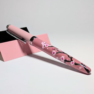 May include: A pink fountain pen featuring a hand-painted black branch design with delicate white and pink cherry blossom flowers. The pen has a silver-toned metal clip and a matching silver band at the center, resting against a pink card.