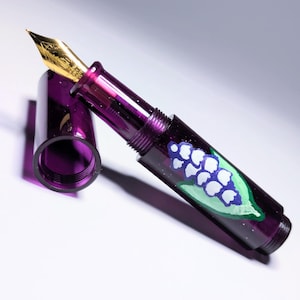 May include: A translucent purple fountain pen with a gold-colored metal nib and a matching purple cap. The barrel of the pen features a hand-painted design of a white flower with green leaves, set against the deep purple resin body of the pen.