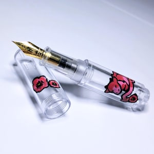 May include: A clear fountain pen with a gold-colored nib and a cap featuring hand-painted red poppy flowers. The barrel of the pen displays a painted red creature with a long tail, also surrounded by small red poppy flower accents.