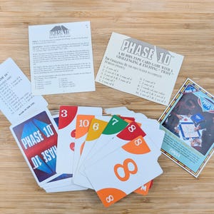 Könnte beinhalten: Ein Phase 10 Kartenspiel-Set mit Anweisungen und Spielkarten. Die Karten haben Zahlen und Farben, mit dem Spielnamen in Blau und Rot. Die Anweisungen sind weiß mit schwarzem Text. Das Spiel befindet sich auf einer Holzoberfläche.