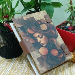 May include: A journal with a brown leather strap closure. The cover features a portrait of a woman with a galaxy-themed design. The journal has a vintage, artistic aesthetic, with a textured paper appearance.