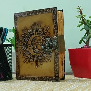 Puede incluir: Un diario de cuero artesanal con un diseño celestial de sol y luna en relieve en la portada. El diario presenta un borde decorativo, un cierre de metal y páginas de papel envejecido. La cubierta es de color marrón dorado cálido.