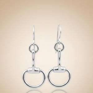 Puede incluir: Un par de pendientes colgantes plateados con temática ecuestre. Cada pendiente presenta un pequeño anillo circular en la parte superior, conectado a una barra vertical y a un diseño de bocado redondeado más grande. Los pendientes se muestran sobre un fondo neutro.