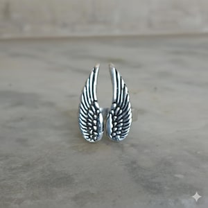 Puede incluir: Un anillo plateado con forma de un par de alas estilizadas. Las alas están detalladas con líneas en forma de plumas, creando una apariencia texturizada. El anillo está sobre un fondo neutro.