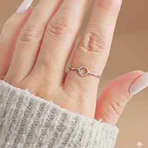 May include: A silver ring featuring a minimalist design with a small, open circle as its centerpiece. The ring is thin and delicate, with a polished finish. The ring is worn on a finger, showcasing its simple elegance. The background is a soft, neutral color.