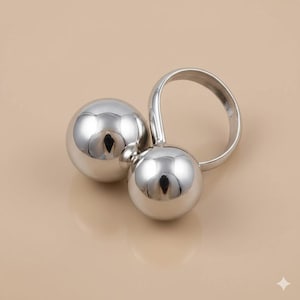 Może przedstawiać: Srebrny pierścionek z dwiema wypolerowanymi, kulistymi kulami połączonymi zakrzywioną opaską. Kule odbijają światło, tworząc jasny, metaliczny wygląd. Pierścionek jest umieszczony na neutralnym tle.