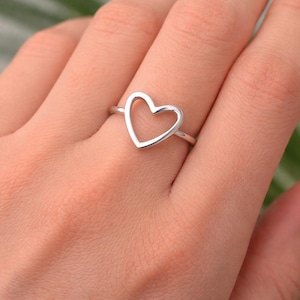 Puede incluir: Un anillo plateado con un diseño en forma de corazón. El corazón es abierto, con una fina banda que forma el contorno. El anillo se lleva en un dedo, mostrando la pieza de joyería simple pero elegante.