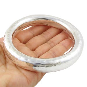 May include: A silver bangle bracelet with a hammered texture. The wide, circular bracelet is held in a hand, showcasing its smooth, reflective surface. The bracelet's design is simple yet elegant, suitable for everyday wear.