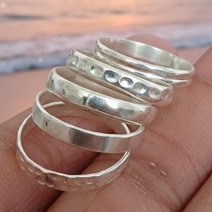 May include: Five silver rings are stacked, each with a distinct design. The rings have different widths and textures, some with hammered or dotted patterns. The rings are shown against a blurred backdrop of a beach and ocean.