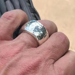 May include: A silver ring with a hammered texture. The ring is thick and has a rounded shape. The metal reflects light, creating highlights and shadows on its surface. The ring is worn on a finger.