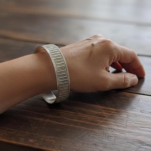 Peut inclure: Un bracelet manchette argenté avec un motif géométrique détaillé. Le bracelet est porté au poignet, mettant en valeur son design. La surface du bracelet est texturée avec de petites lignes et points espacés, créant un rythme visuel.