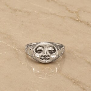 Könnte beinhalten: Ein silberfarbener Metallring mit einem erhabenen, skulptierten Mondgesicht mit geschlossenen Augen und einem lächelnden Mund. Das Ringband ist mit kleinen, geprägten Sternformen verziert, die sich vom zentralen Mondgesicht zu den Seiten erstrecken.