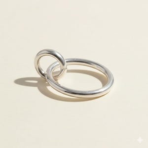 Peut inclure: Une bague argentée avec un design en forme de lien. La bague présente un anneau circulaire plus grand relié à un anneau circulaire plus petit. Le métal a une surface polie et réfléchissante, sur fond neutre.