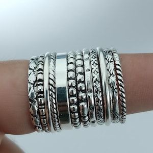 May include: A collection of silver rings with various textures and designs. The rings include smooth bands, beaded patterns, and detailed floral and geometric motifs. The rings are stacked together, showcasing a variety of styles.