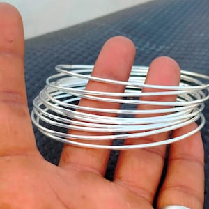 May include: A stack of silver-colored bangles. The thin, round bracelets are held in a hand, with several visible on the fingers and palm. The bangles are made of a shiny, metallic material.