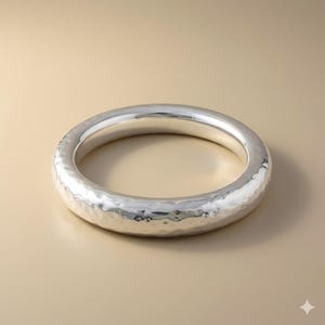 May include: A silver-colored, hammered metal bangle bracelet. The bracelet has a thick, rounded design with a textured surface. The bangle is circular and appears to be made of a shiny, reflective metal. The background is a solid, neutral color.