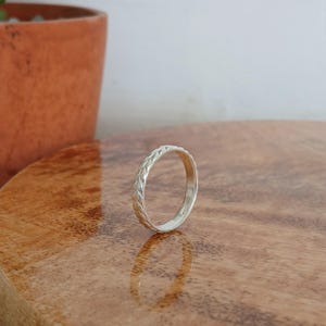 Puede incluir: Un anillo de plata con un diseño trenzado detallado. El anillo está sobre una superficie de madera, con un fondo borroso que presenta una maceta de terracota y una pared blanca. El intrincado patrón del anillo es claramente visible.