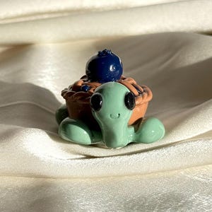 May include: A whimsical, handmade turtle figurine with a light green body and a pie-shaped shell. The shell is brown with a blueberry on top. The turtle has large, black eyes and is set against a white background.