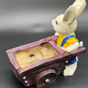 Könnte beinhalten: Eine Keramik-Hasenfigur, die einen kleinen Wagen im rustikalen Stil schiebt. Der Hase trägt ein gelb-blaues Outfit. Der Wagen ist braun mit hellem Innenraum, und die Räder sind sichtbar.