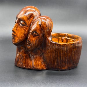 May include: A brown ceramic planter with two sculpted faces. The faces have closed eyes and textured surfaces. The planter has a rectangular shape with a hollowed-out area for plants or decorative items. The design is artistic and unique, suitable for home decor.