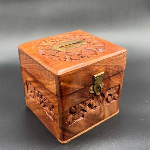 Op de afbeelding: Een vierkante houten doos met ingewikkelde bloemsnijwerken en een messing sluiting. De doos is gemaakt van roodbruin hout en heeft een sleuf aan de bovenkant. De doos wordt waarschijnlijk gebruikt voor het opbergen van kleine items of als decoratief stuk.