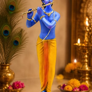 May include: A blue-skinned figurine of a deity standing on a dark base, playing a gold-coloured flute. The figure wears a yellow dhoti, a gold crown, and gold jewellery, including a necklace and armbands, with a serene expression and closed eyes.