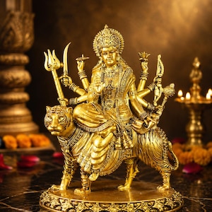 Könnte beinhalten: Eine goldfarbene Statue einer vielarmigen Gottheit, die auf einem Tiger sitzt. Die Figur trägt eine verzierte Krone und Schmuck und hält in jeder Hand verschiedene symbolische Gegenstände, darunter einen Dreizack und ein Schwert.
