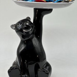 Peut inclure: Un plat de service en céramique noir en forme d'ours, avec une assiette blanche au-dessus de sa tête. L'assiette est remplie de bonbons emballés colorés. L'ours est brillant et a une expression souriante.