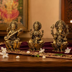 Puede incluir: Tres estatuas de metal en tono dorado que representan deidades sentadas sobre pedestales de loto. La figura de la izquierda sostiene un instrumento de cuerda, la central tiene varios brazos en gesto de bendición y la derecha tiene cabeza de elefante.