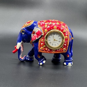 Puede incluir: Figura decorativa de elefante en azul, rojo y dorado, con un reloj en un lateral. El elefante tiene patrones detallados y una esfera dorada con números romanos negros. Un artículo de decoración del hogar único.