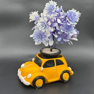 Może przedstawiać: Żółta doniczka w kształcie samochodu z czarnymi kołami i oknami, trzymająca małą doniczkę ze sztucznymi fioletowymi i białymi kwiatami. Doniczka ma styl vintage.