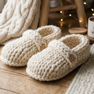 May include: A pair of cream-colored, chunky knit slippers with wooden button details, resting on a wooden floor. A matching knit blanket and a mug with "COZY WINTER" text are also visible, suggesting a theme of comfort and warmth.