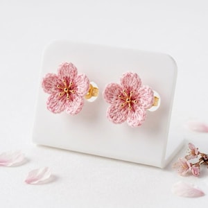 May include: A pair of stud earrings featuring five-petaled pink crocheted flowers. Each flower has a gold-toned metal center with thin, radiating stamens. The earrings are displayed on a white stand, accompanied by loose pink flower petals.