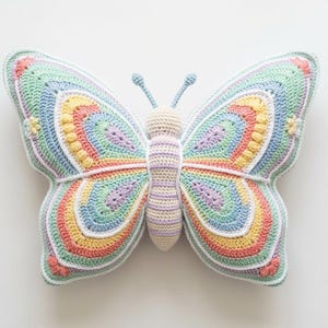 Puede incluir: Un cojín de mariposa de ganchillo colorido con un diseño de arcoíris. Las alas presentan círculos concéntricos de colores pastel, incluyendo azul, amarillo y rosa. El cuerpo es de color crema y las antenas son de color azul claro.