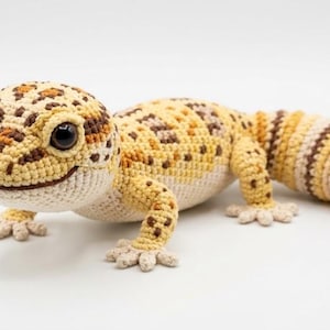 May include: A crocheted leopard gecko in shades of yellow, brown, and cream. The handmade reptile features a detailed pattern, including spots on its head and back, and is posed on a white surface.