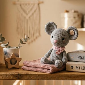 Pode incluir: Um rato cinzento de crochê com um cachecol rosa está sentado em uma manta rosa. Ao lado, dois livros com os títulos "NIGHT BEAR" e "MY LITTLE WORLD". Uma estrela de madeira e um vaso com vegetação também estão na prateleira.