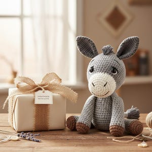 May include: A handmade gray crochet donkey with a white muzzle and brown hooves sits next to a wrapped gift box. The gift box is tied with a burlap ribbon and a tag that reads "HANDMADE WITH LOVE".