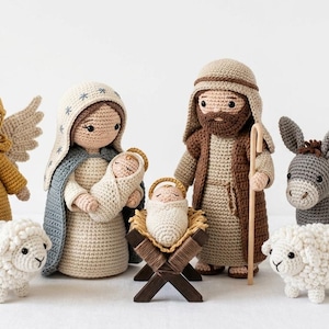 Op de afbeelding: Een gehaakte kerststal met figuren van een engel, Maria, Jozef, baby Jezus in een kribbe, twee schapen, een ezel en een koe. De figuren zijn in verschillende kleuren, waaronder geel, wit, bruin en grijs.