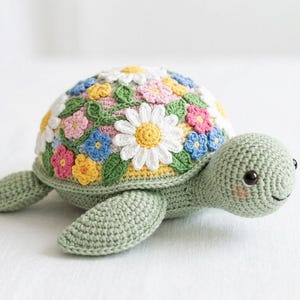 May include: A handmade crochet turtle with a floral shell design. The turtle's shell is adorned with colorful crochet flowers in white, pink, blue, and yellow. The turtle has a light green body and a smiling face.