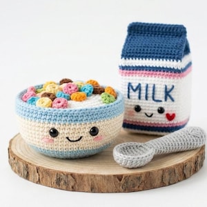 May include: A crocheted breakfast set featuring a bowl of colorful cereal, a milk carton with the word "MILK", and a spoon. The bowl and milk carton have smiling faces. The set is displayed on a wooden slice.