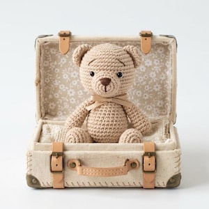 May include: A crocheted teddy bear in a vintage-style suitcase. The bear is light brown with a matching bow. The suitcase is beige with leather straps and a floral interior lining. A charming, handmade item.