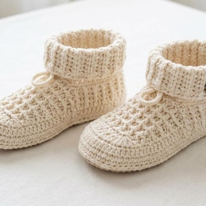 Könnte beinhalten: Ein Paar cremefarbene, handgefertigte Häkelschuhe. Die Schuhe haben ein strukturiertes Muster am Fuß und einen gerippten Bund, der umgeschlagen werden kann. Jeder Schuh hat einen Knopf- und Schlaufenverschluss.